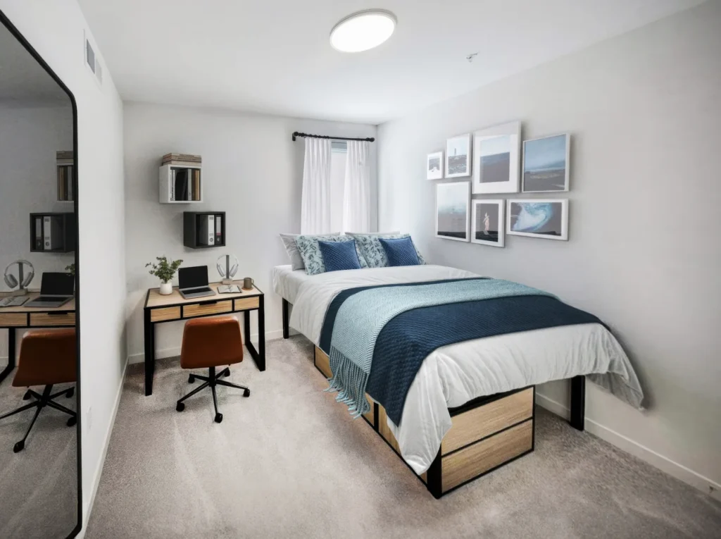 Bedroom with window, mounted wall mirror, desks, chair, laptop, and multiple pictures hanging on the wall in view.
