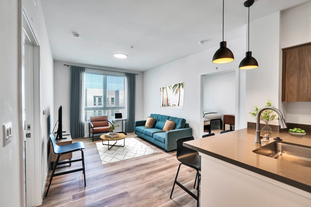 Living Room with desks, chair, tv, laptop, bed, tropical wall painting, and kitchen sink in view.