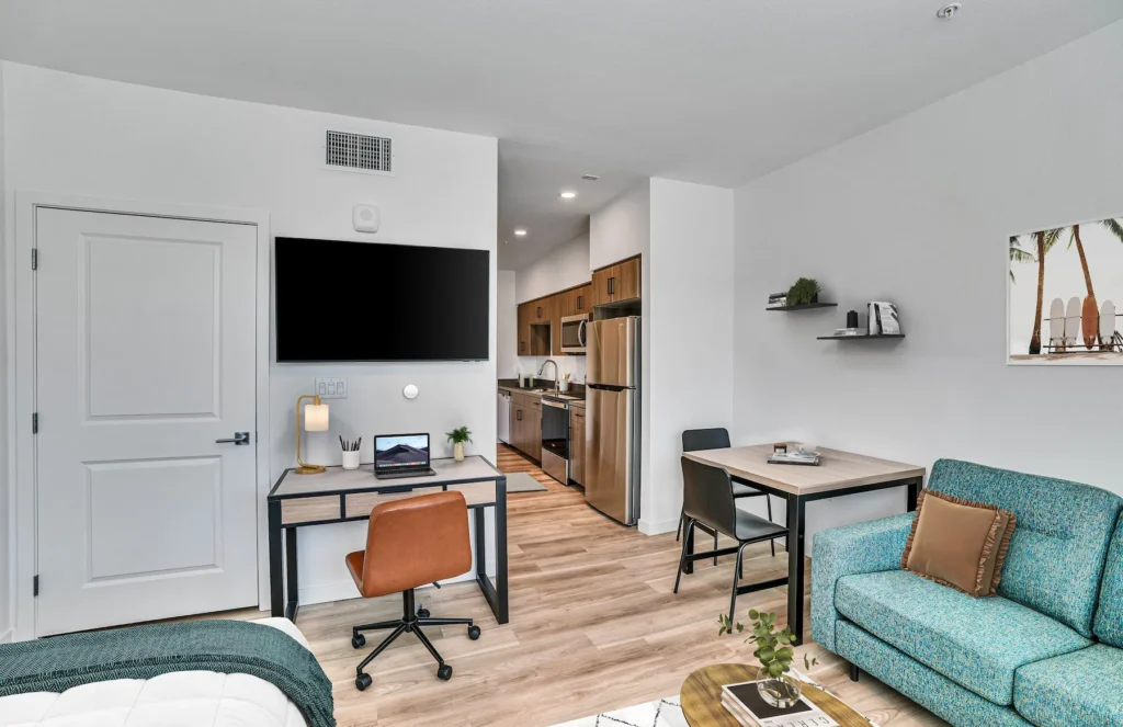 Living Room with desks, chair, wall mounted tv, laptop, bed, tropical wall painting, and kitchen in view.